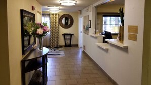Un espace d’accueil d’hôtel comprenant un comptoir de réception, un miroir, un vase avec des fleurs, un rideau à motifs et une horloge murale.