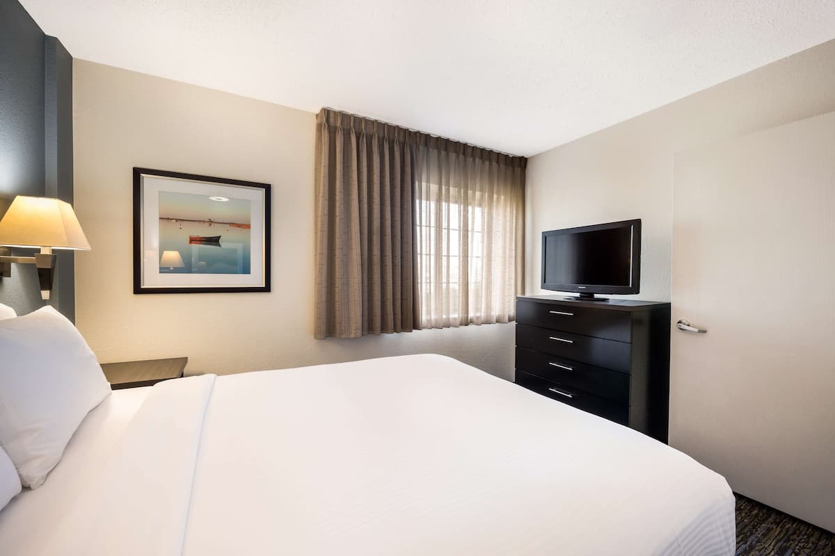 Een hotelkamer met een groot bed, een televisie op een dressoir, een ingelijst schilderij aan de muur en een raam met gordijnen.