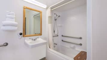 Un baño moderno con un lavamanos blanco, un espejo, una ducha con barras de apoyo y un tapete de baño de madera.