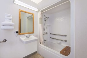 Un baño moderno con un lavamanos blanco, un espejo, una ducha con barras de apoyo y un tapete de baño de madera.