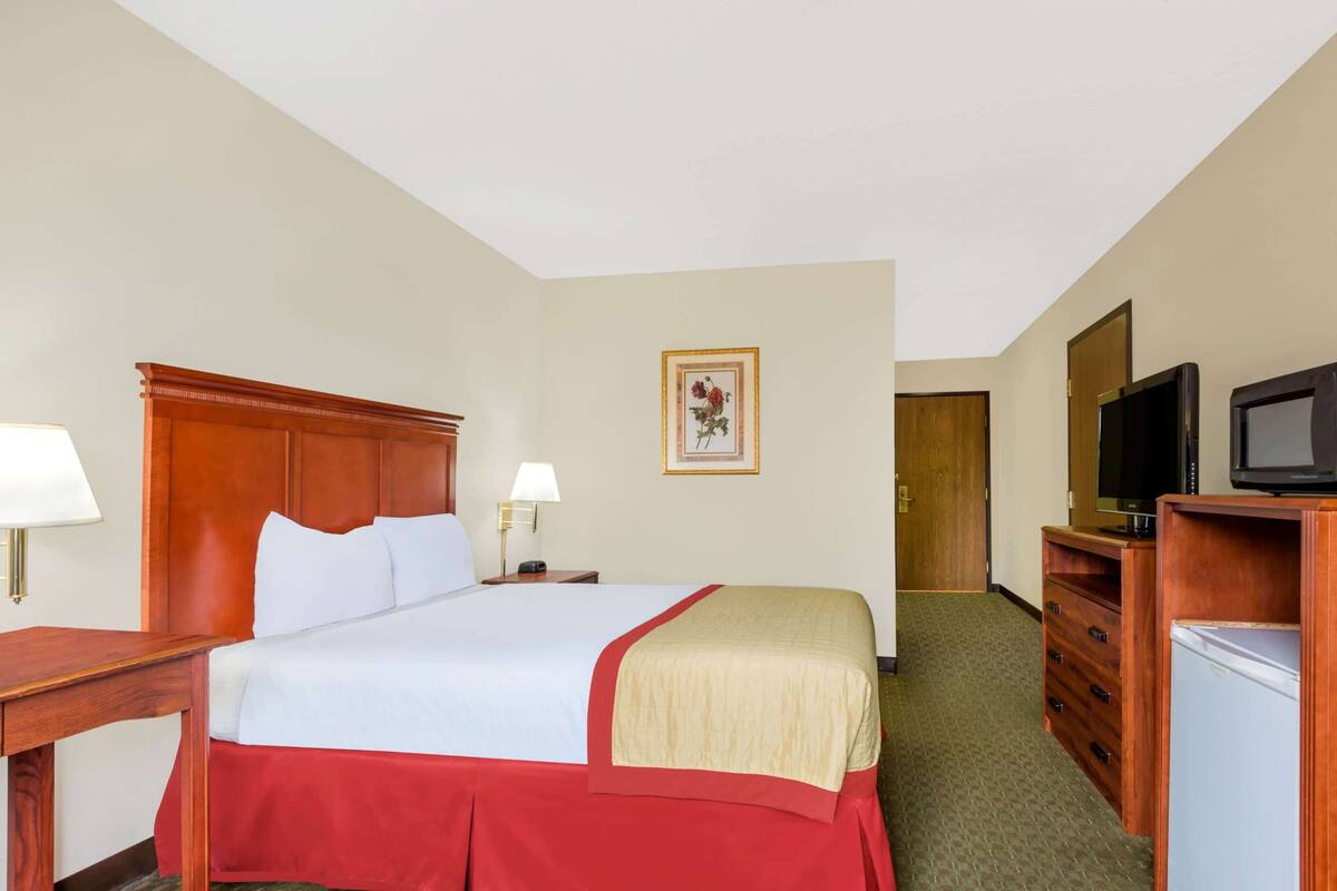 A hotel room with a bed, bedside tables, a television, a refrigerator, and a door leading to another room.