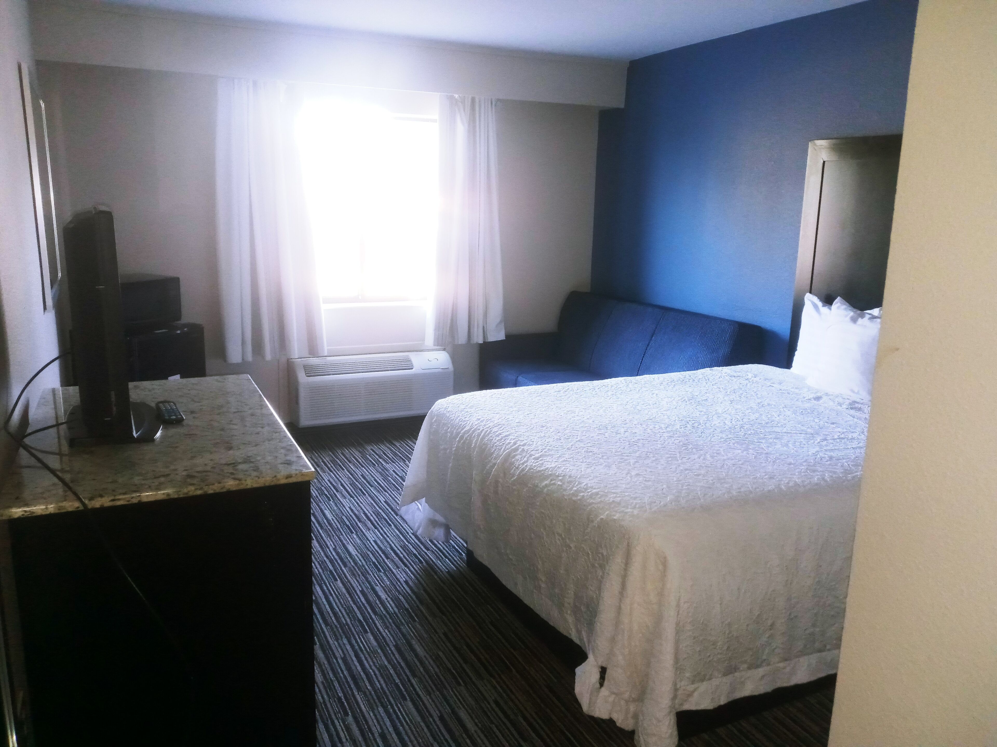 Una habitación de hotel con una cama, un escritorio con una computadora y un control remoto, una ventana con cortinas, un aire acondicionado y una pared azul.