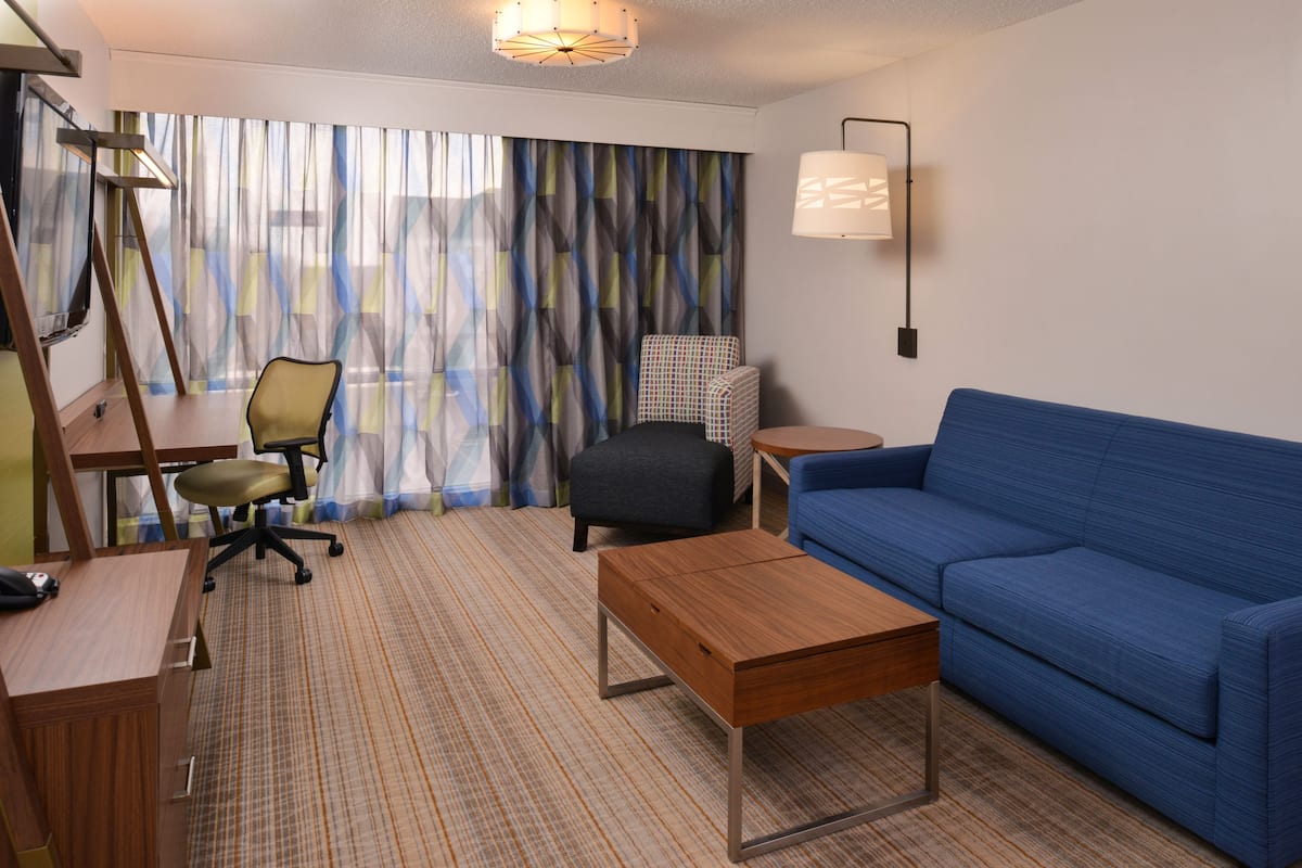 A hotel room with a blue sofa, a wooden desk, a chair, a small table, a lamp, and a window with curtains.