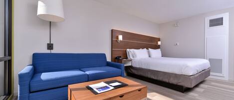 A modern hotel room with a blue sofa, a wooden coffee table, a bed with white bedding, and a wall-mounted lamp.