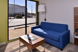 Una habitación moderna con un sofá azul, una mesa de centro de madera y un amplio ventanal con vistas a un estacionamiento y un edificio.