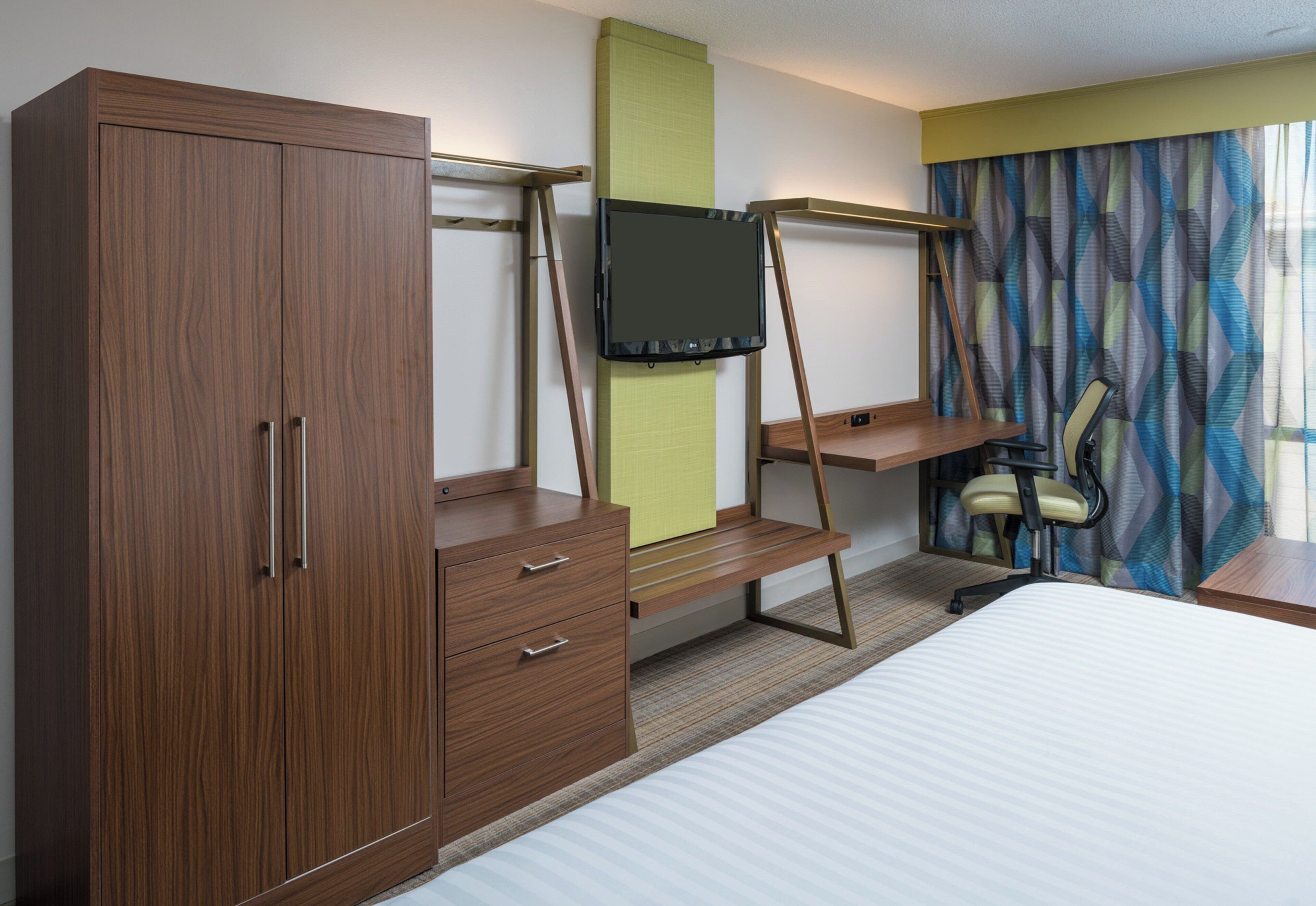 A hotel room with a bed, a wooden wardrobe, a desk with a television, and a chair.