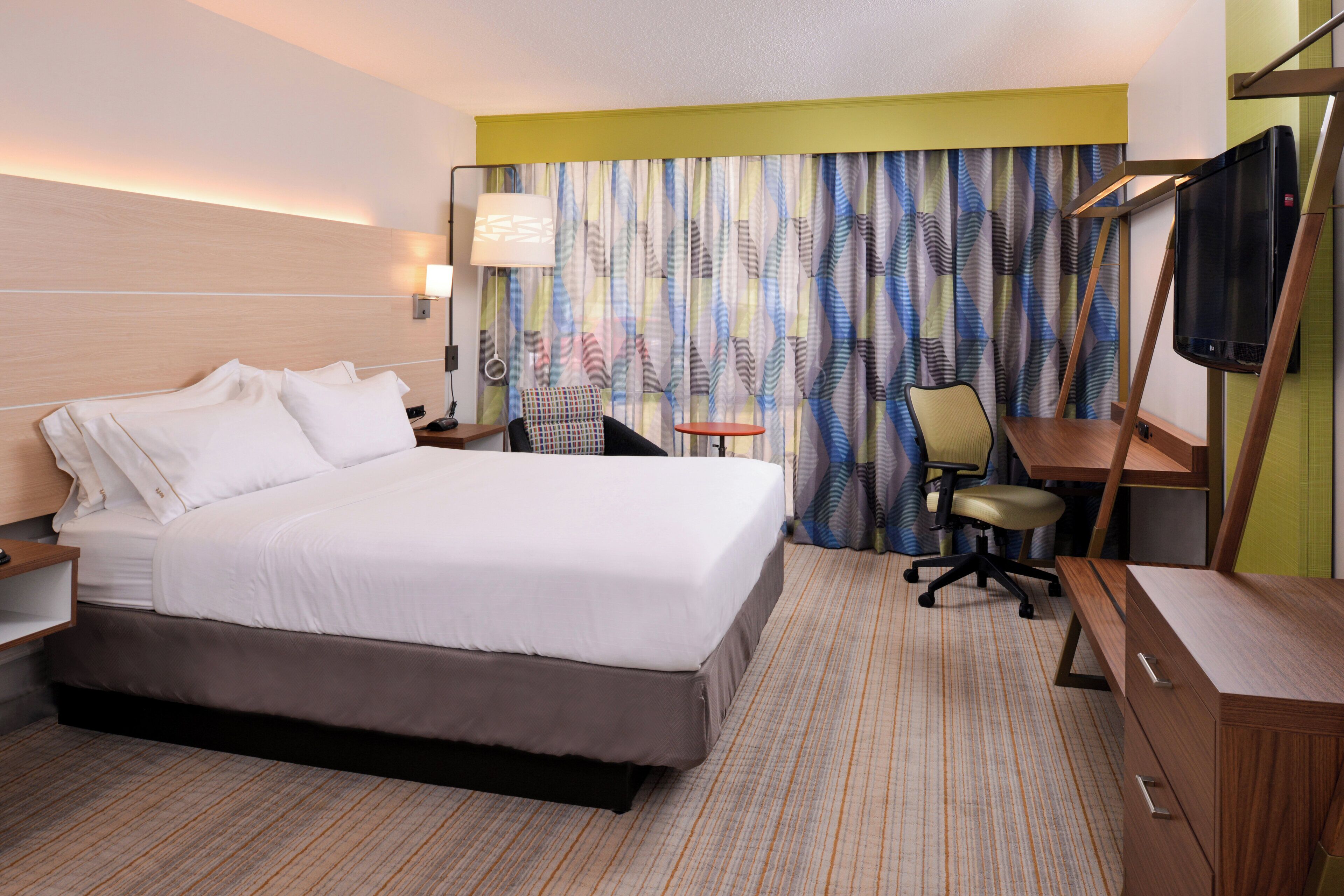 A hotel room with a large bed, a desk, a chair, a television, and a window with curtains.