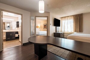 Quarto de hotel com uma cama grande, uma mesa de trabalho, televisão de tela plana, banheiro com espelho e pia, e vista para o exterior.