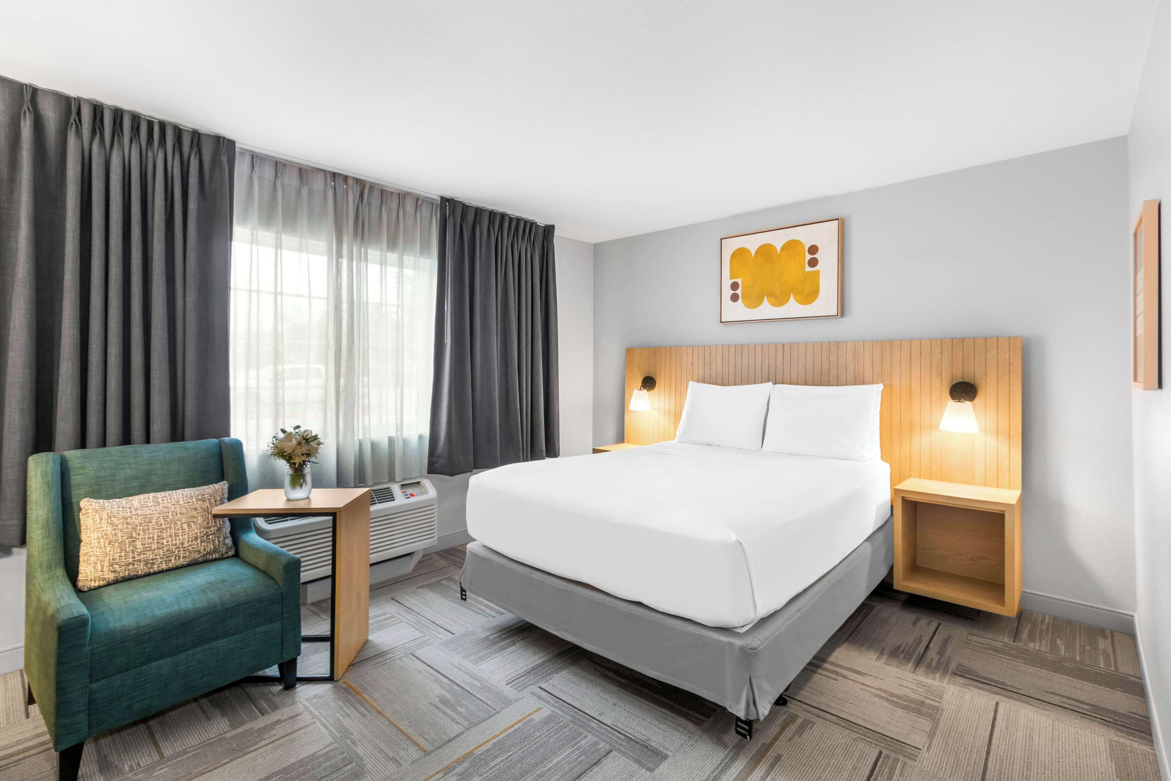 A hotel room with a bed, a green armchair, a small table with a vase, and a wall-mounted artwork.