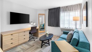 A modern hotel room with a flat-screen TV, a wooden dresser, a blue sofa, and a small table with a vase of flowers. - Sonesta Simply Suites Austin South (Austin)