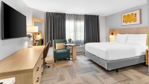 A hotel room with a bed, a desk, a chair, a TV, a lamp, a small table with a vase, and a window with curtains. - Sonesta Simply Suites Austin South (Austin)