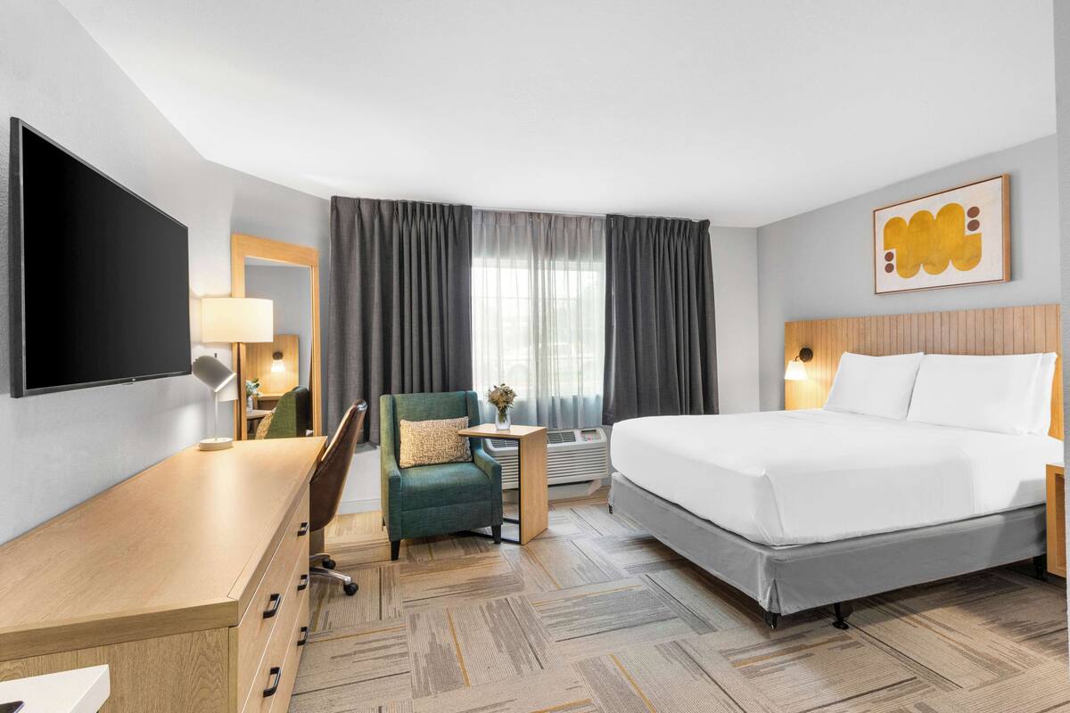 Una habitación de hotel con cama, escritorio, silla, televisor, lámpara, una mesita con un florero y una ventana con cortinas.