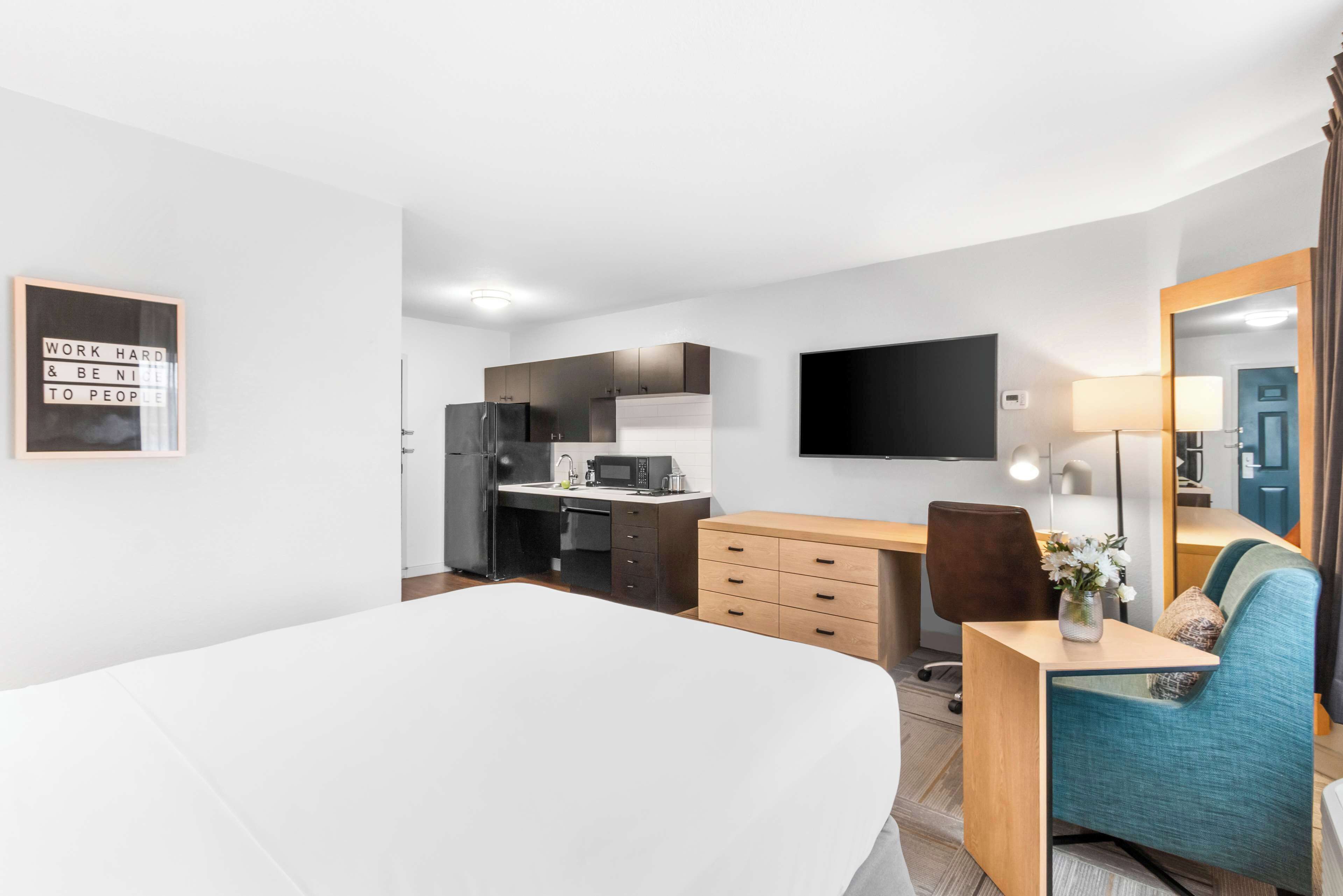 Una habitación de hotel moderna con una cama grande, un escritorio con una silla, una kitchenette y un televisor montado en la pared.
