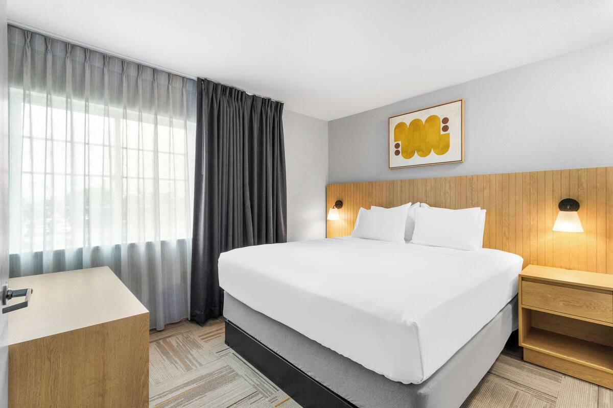 Una habitación de hotel con una cama grande, una mesita de noche, una lámpara de pared y un ventanal con cortinas.