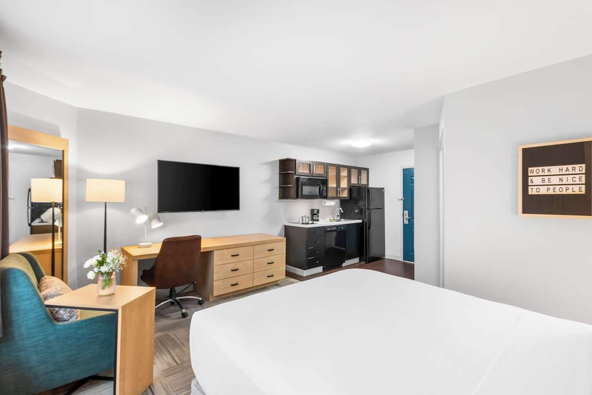 Habitación de hotel con cama, escritorio con silla, kitchenette pequeña y televisor montado en la pared.