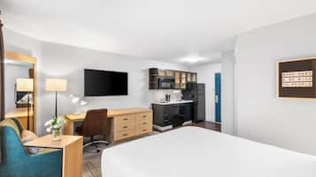 A hotel room with a bed, a desk with a chair, a small kitchenette, and a TV mounted on the wall.