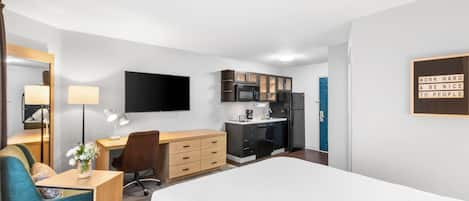 A hotel room with a bed, a desk with a chair, a small kitchenette, and a TV mounted on the wall.