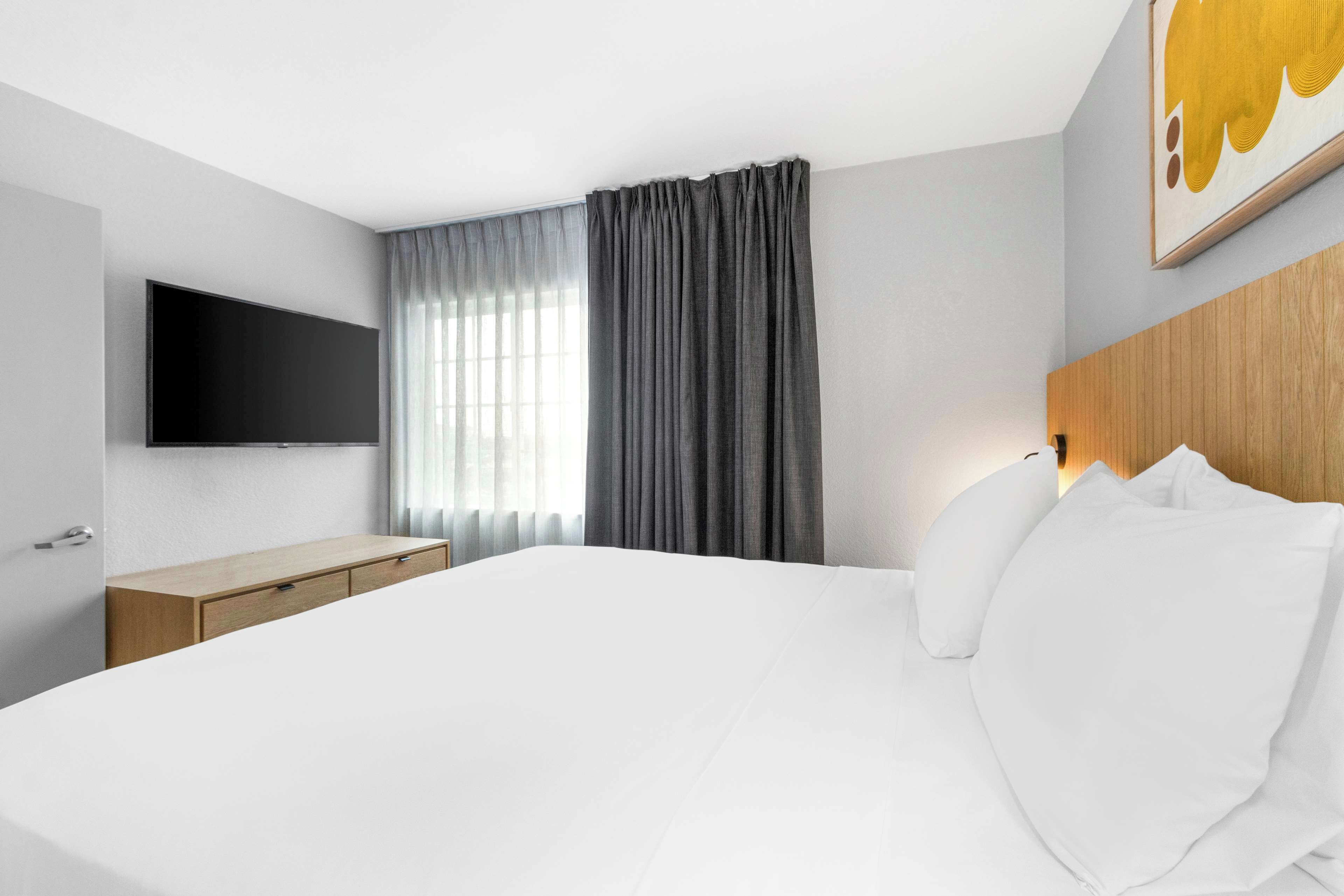 A modern hotel room with a large bed, a flat-screen TV mounted on the wall, a wooden nightstand, and a window with grey curtains.