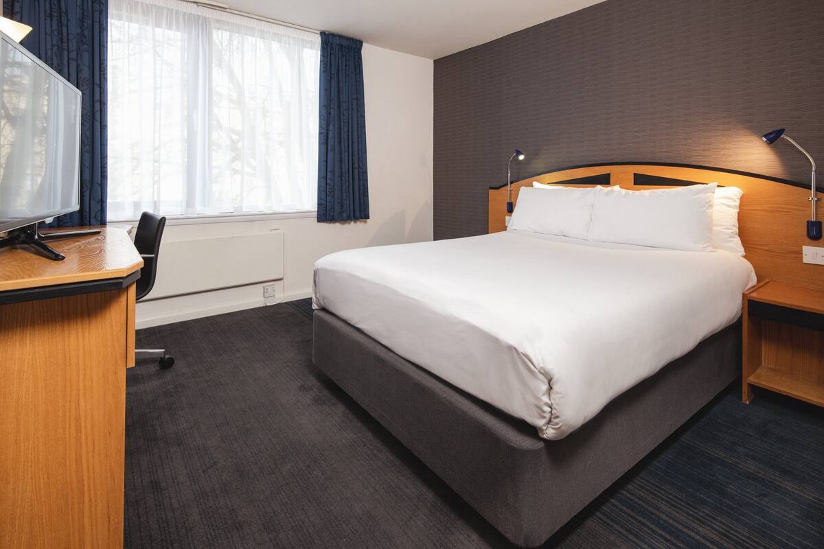A hotel room with a large bed, a desk with a computer, a chair, and a window with curtains.
