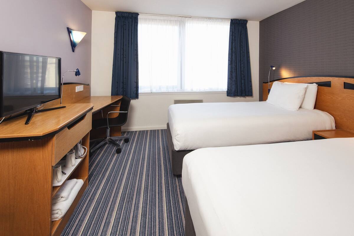 A hotel room with two beds, a desk with a TV, a chair, and a window with curtains.