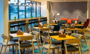 A modern restaurant with multiple seating areas, including tables and chairs, and a large window offering a view of the city. - Holiday Inn Express Bristol City Centre by IHG (Bristol)