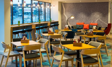 A modern restaurant with multiple seating areas, including tables and chairs, and a large window offering a view of the city.