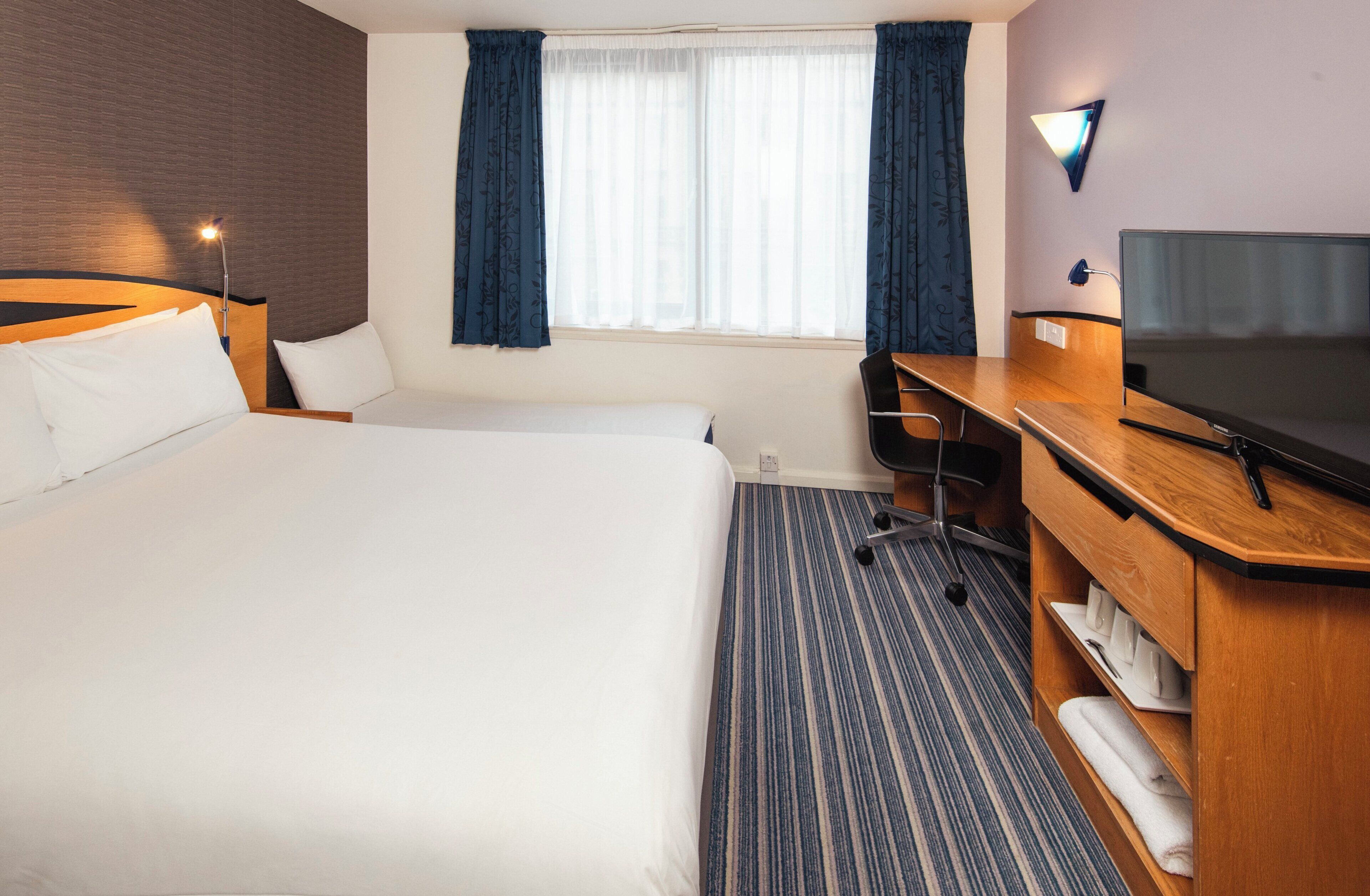 A hotel room with a large bed, a desk, a chair, a television, and a window with curtains.