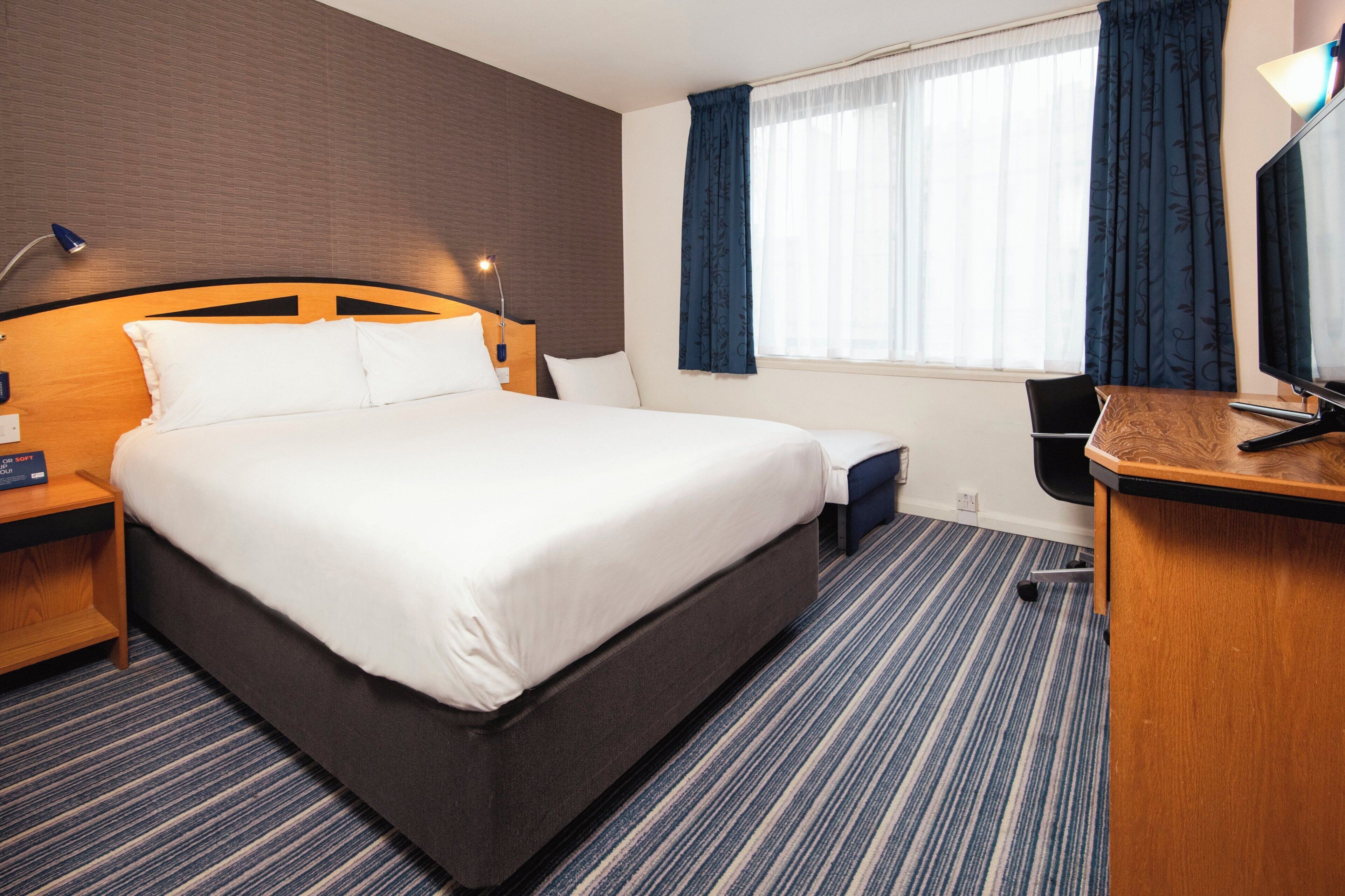 Une chambre d’hôtel avec un grand lit, un bureau avec une chaise, une lampe et une fenêtre avec des rideaux.