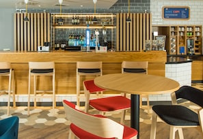 Zona de bar con mostrador de madera, taburetes y una mesa redonda con sillas. Hay estantes con botellas y un letrero sobre la barra.