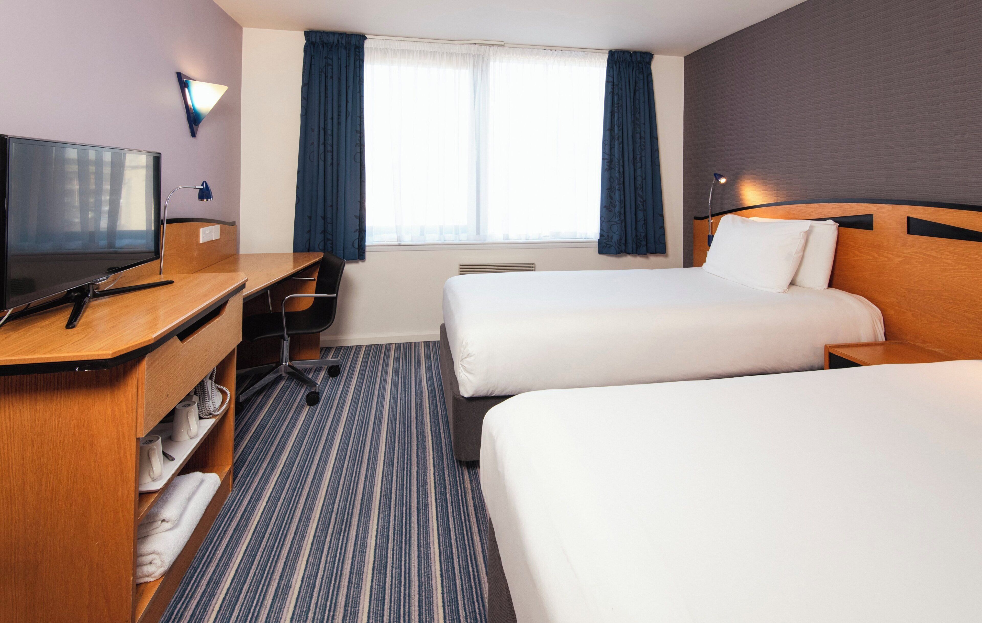 A hotel room with two beds, a desk with a TV, and a window with curtains.