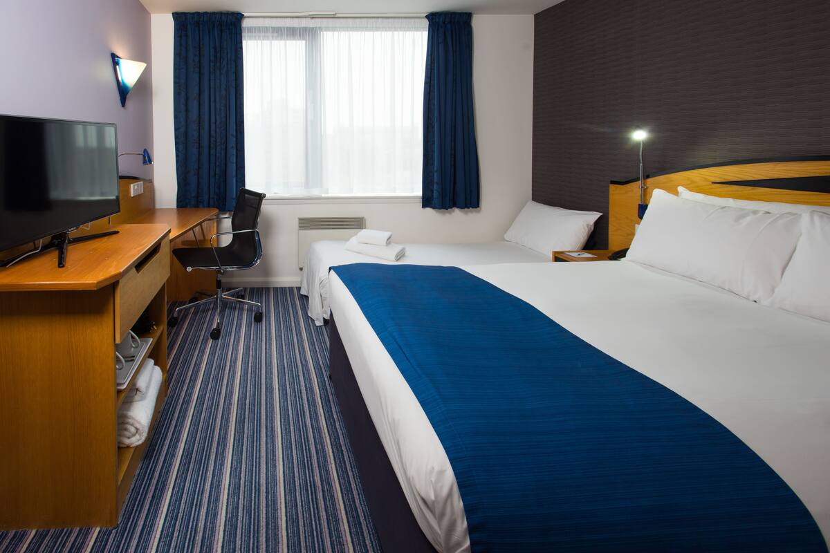 A hotel room with a bed, a desk with a TV, a chair, a window with curtains, and a bedside lamp.