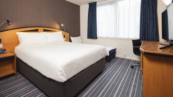 Quarto de hotel com uma cama grande, uma escrivaninha com computador, uma cadeira, um abajur e uma janela com cortinas.