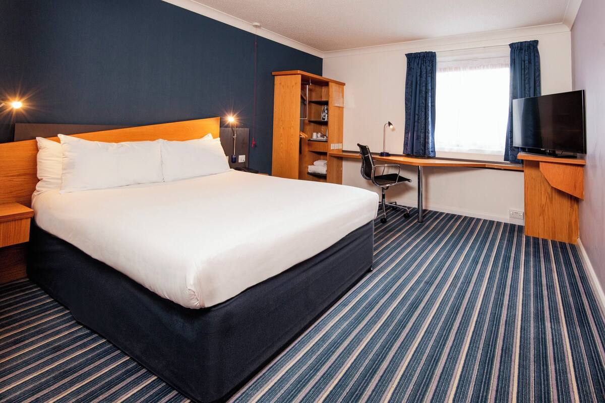 A hotel room with a large bed, a desk with a chair, a television, and a wardrobe.