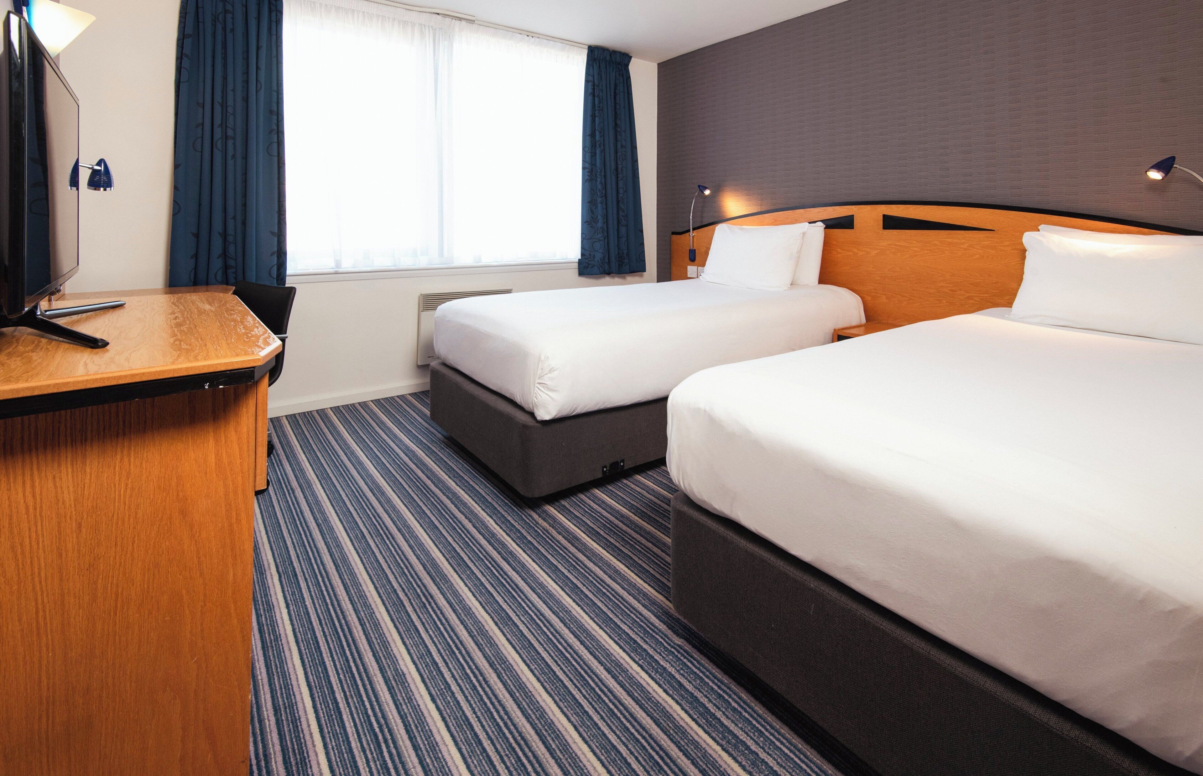Ein Hotelzimmer mit zwei Betten, einem Schreibtisch, einer Lampe und einem Fenster mit Vorhängen.