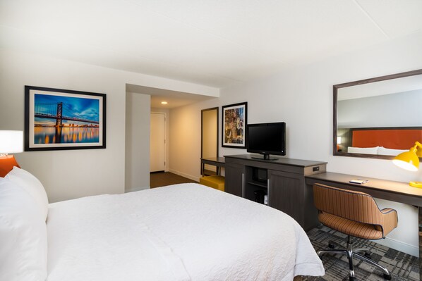 Quarto de hotel com cama, escrivaninha, cadeira, televisão e obras de arte nas paredes.