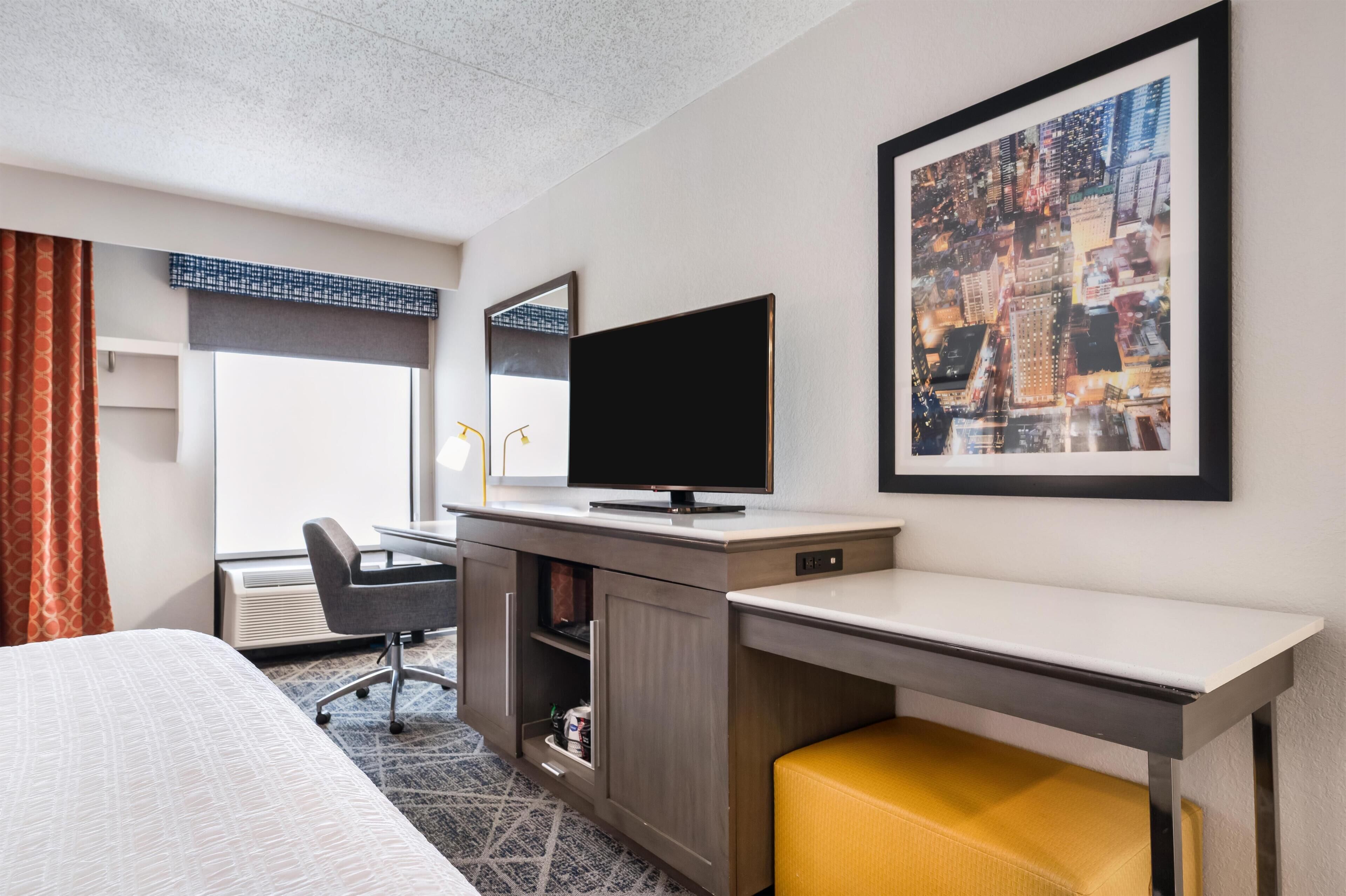 Quarto de hotel com cama, escrivaninha com computador, cadeira, televisão, quadro grande emoldurado e janela com cortinas.