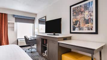 Quarto de hotel com cama, escrivaninha com computador, cadeira, televisão, quadro grande emoldurado e janela com cortinas.