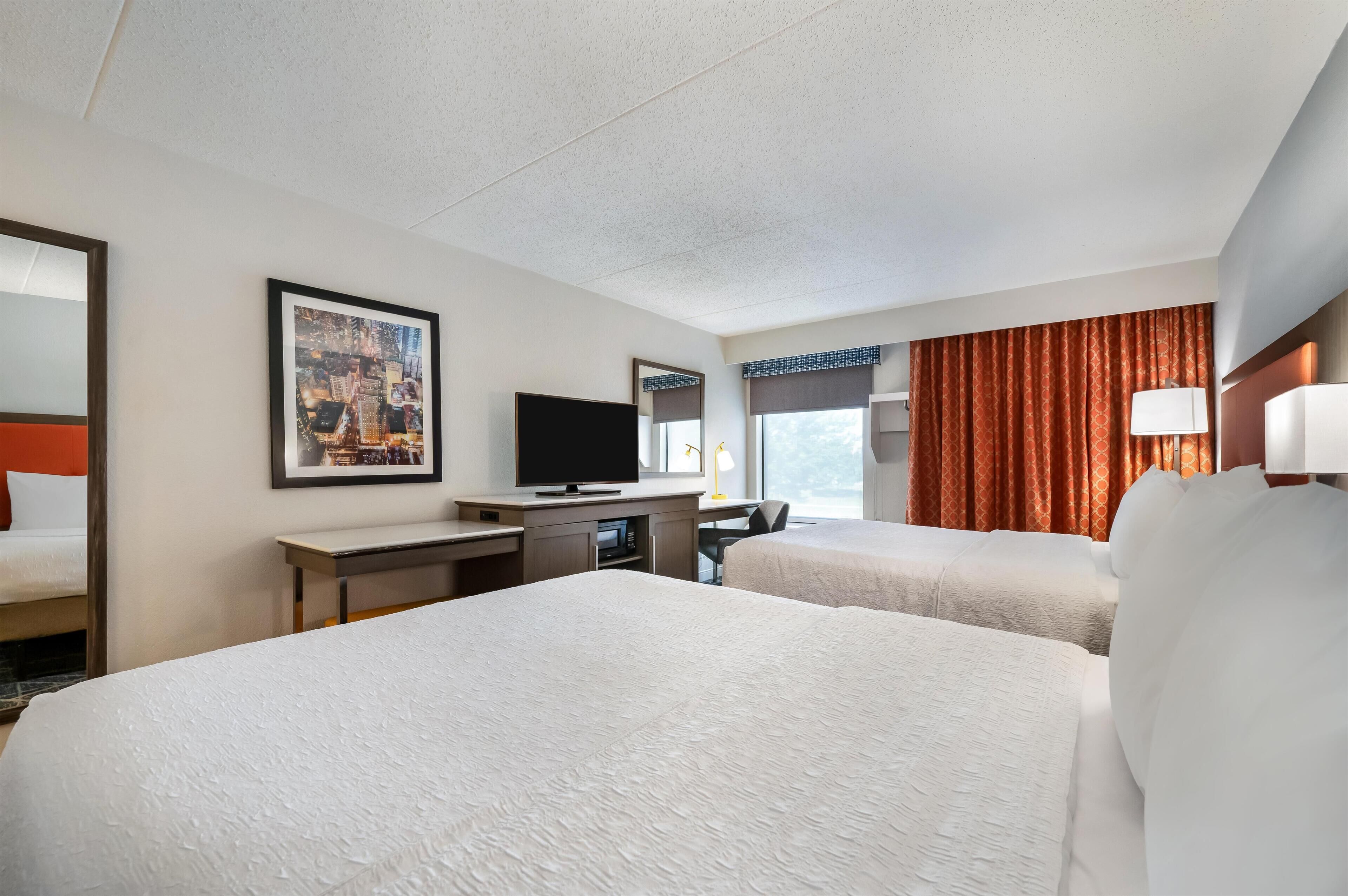 A hotel room with two beds, a desk with a TV, a window with curtains, and a framed picture on the wall.