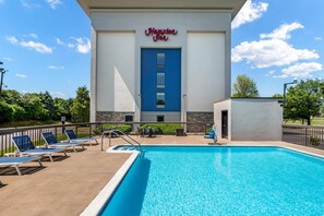 Une piscine avec des chaises longues, une enseigne « Hampton Inn » et un ciel bleu azur.