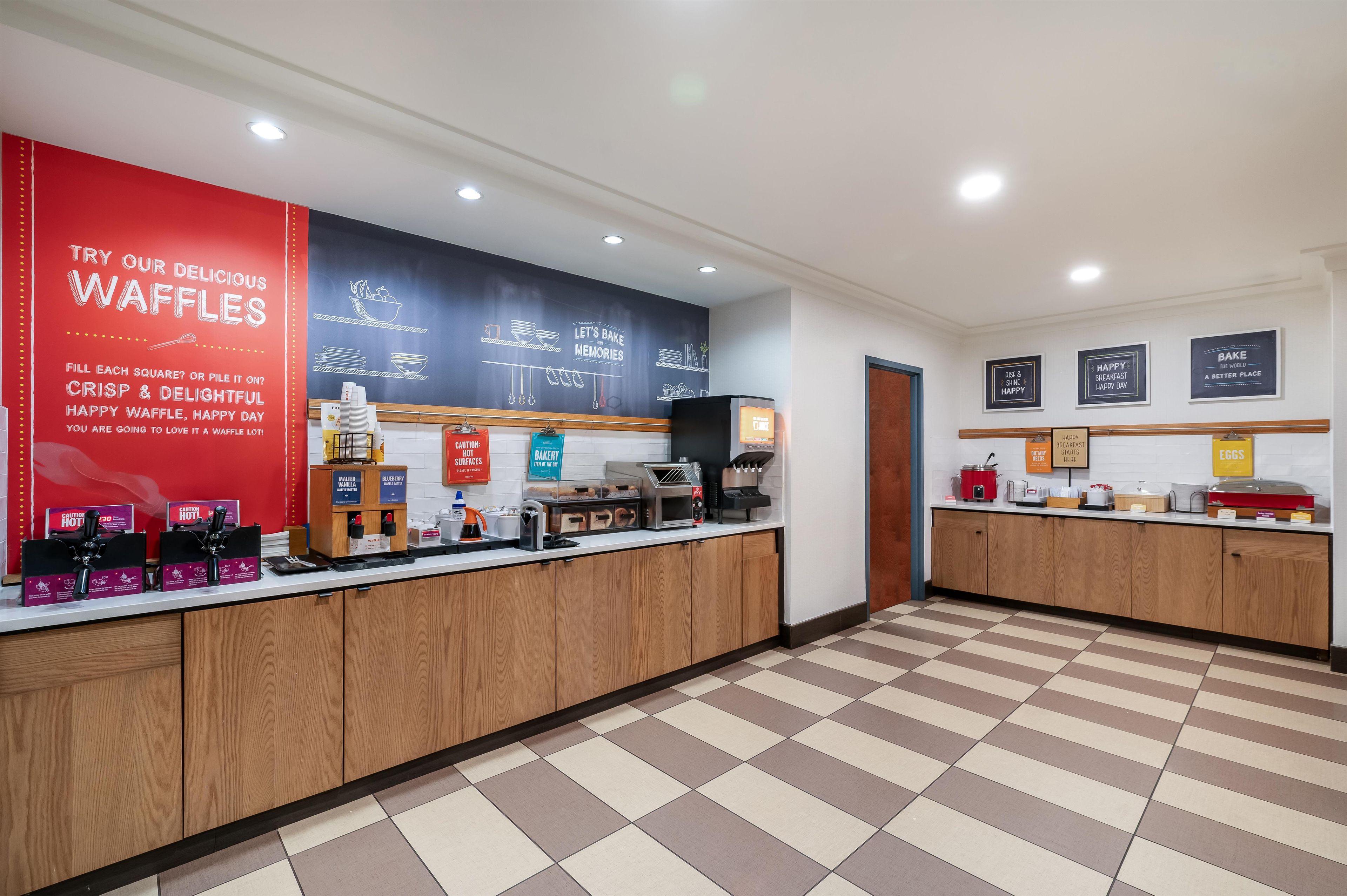 Mostrador de panadería con equipos para hacer gofres, un letrero rojo que promociona los gofres y una pared con decoración relacionada con la repostería.