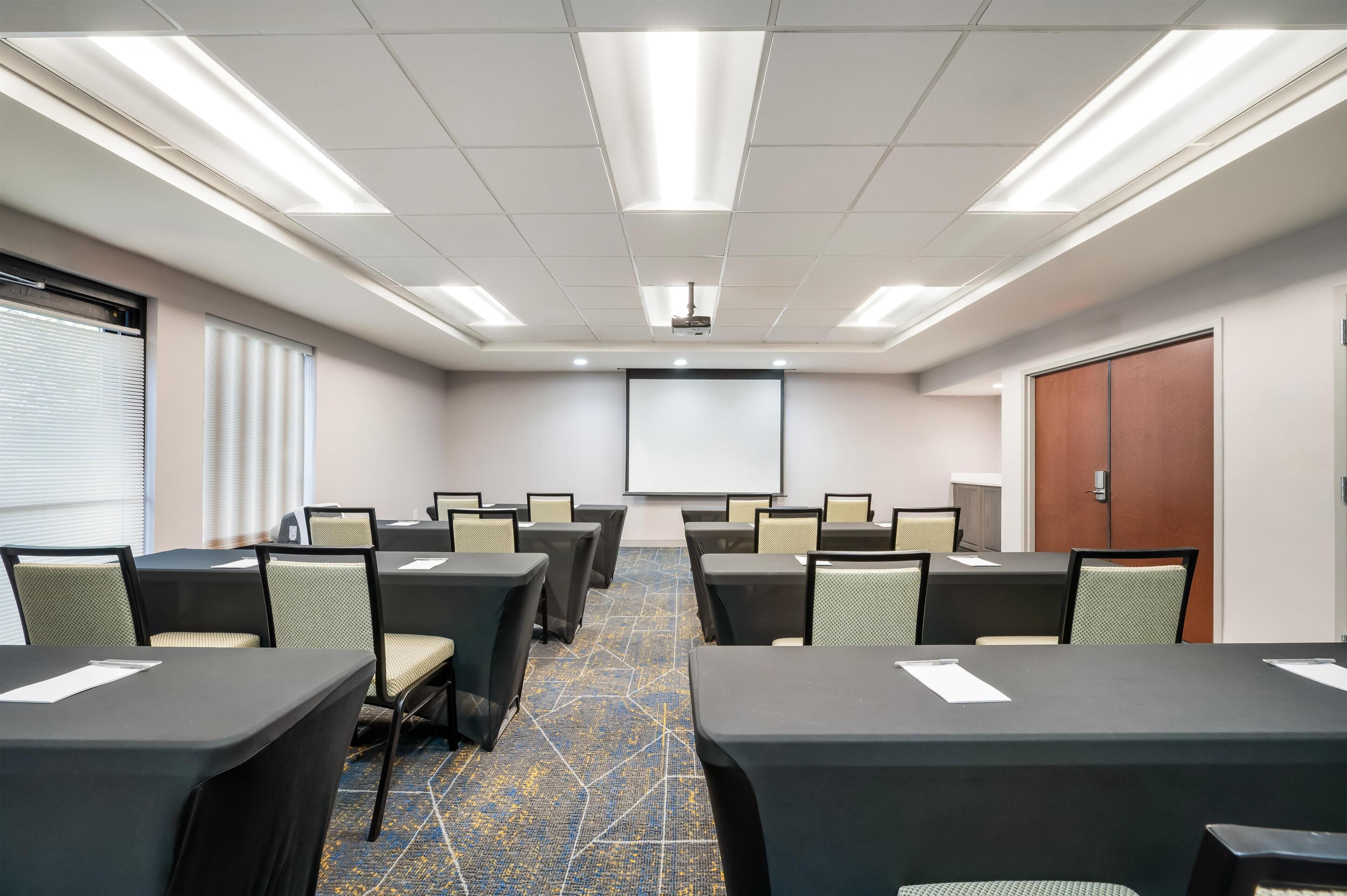 Une salle de conférence équipée de tables, de chaises, d’un écran de projection et d’une porte.