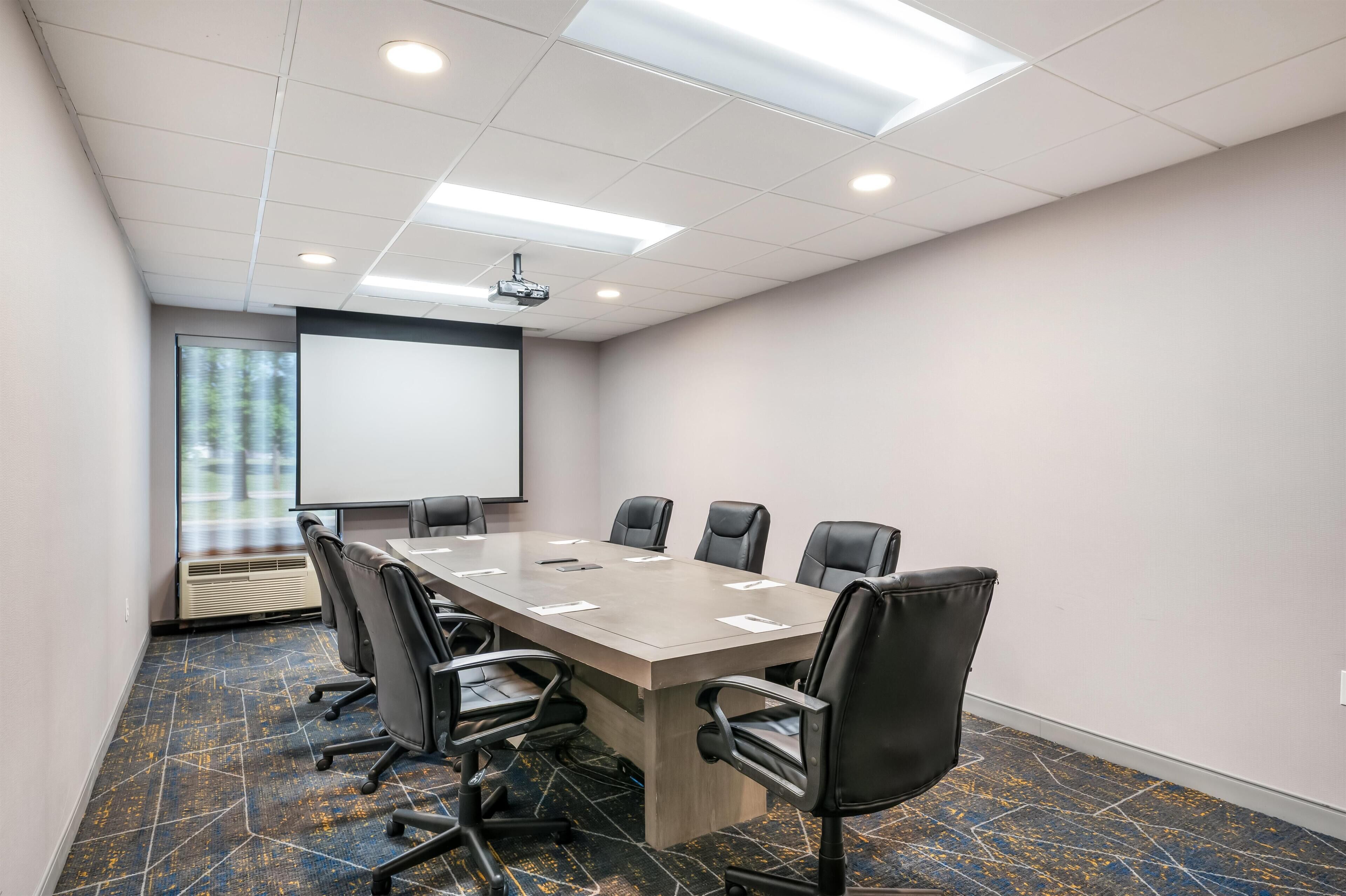 A conference room with a long table, black chairs, a projector screen, and a large window.