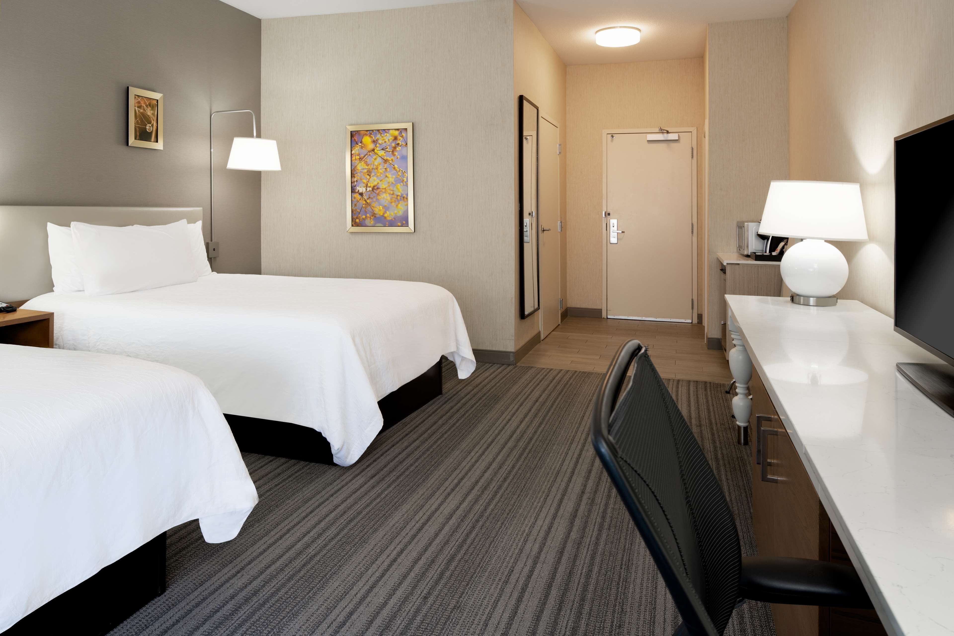A hotel room with two beds, a desk, a chair, a TV, a lamp, and a door leading to a bathroom.