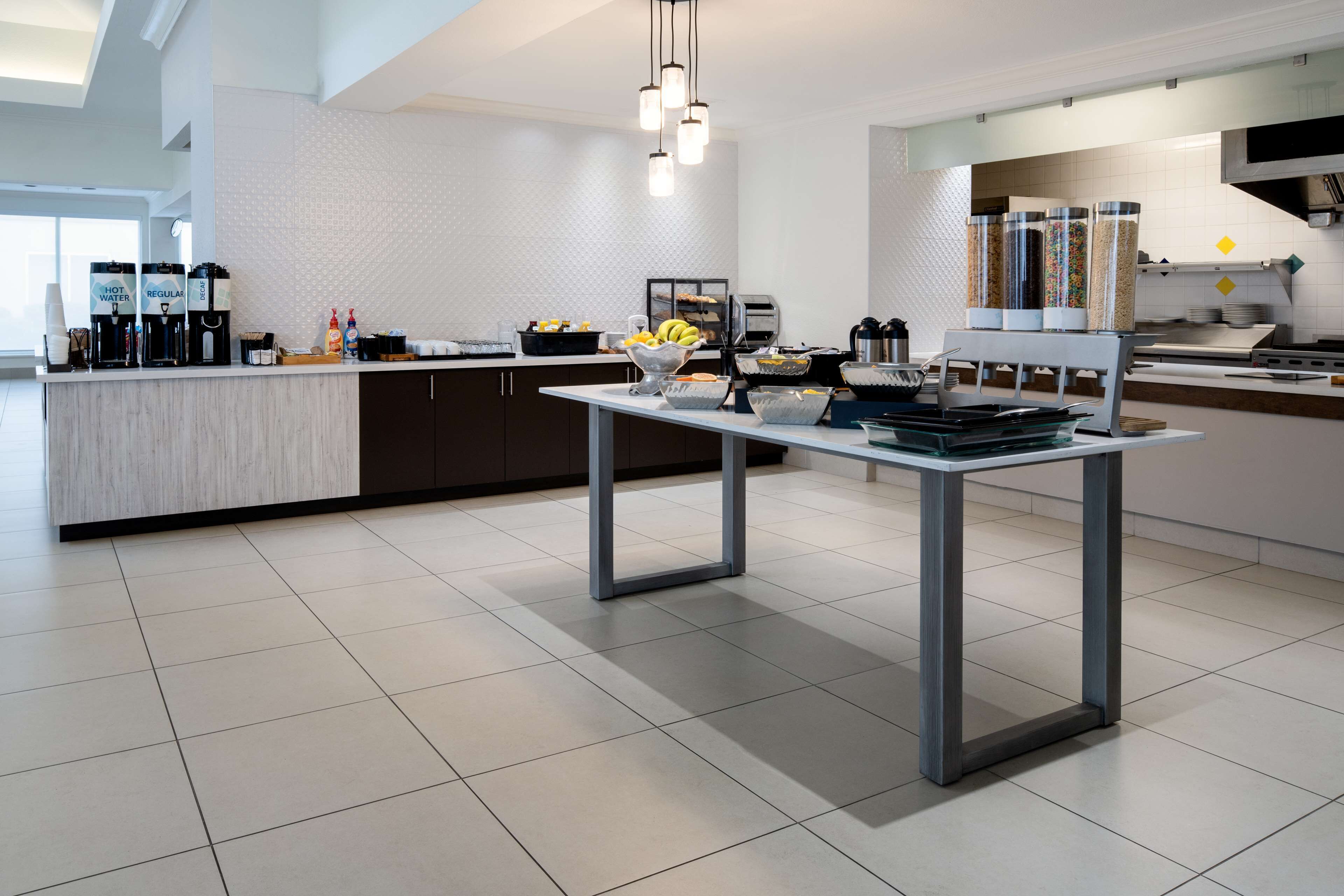A modern kitchen with a central island, coffee machines, and a variety of food items.