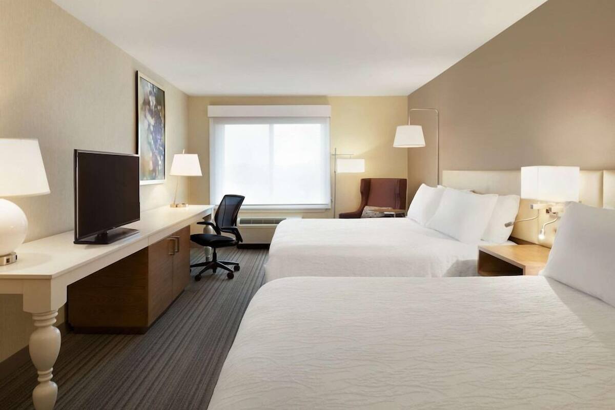 A hotel room with two beds, a desk with a TV, a chair, and a window.