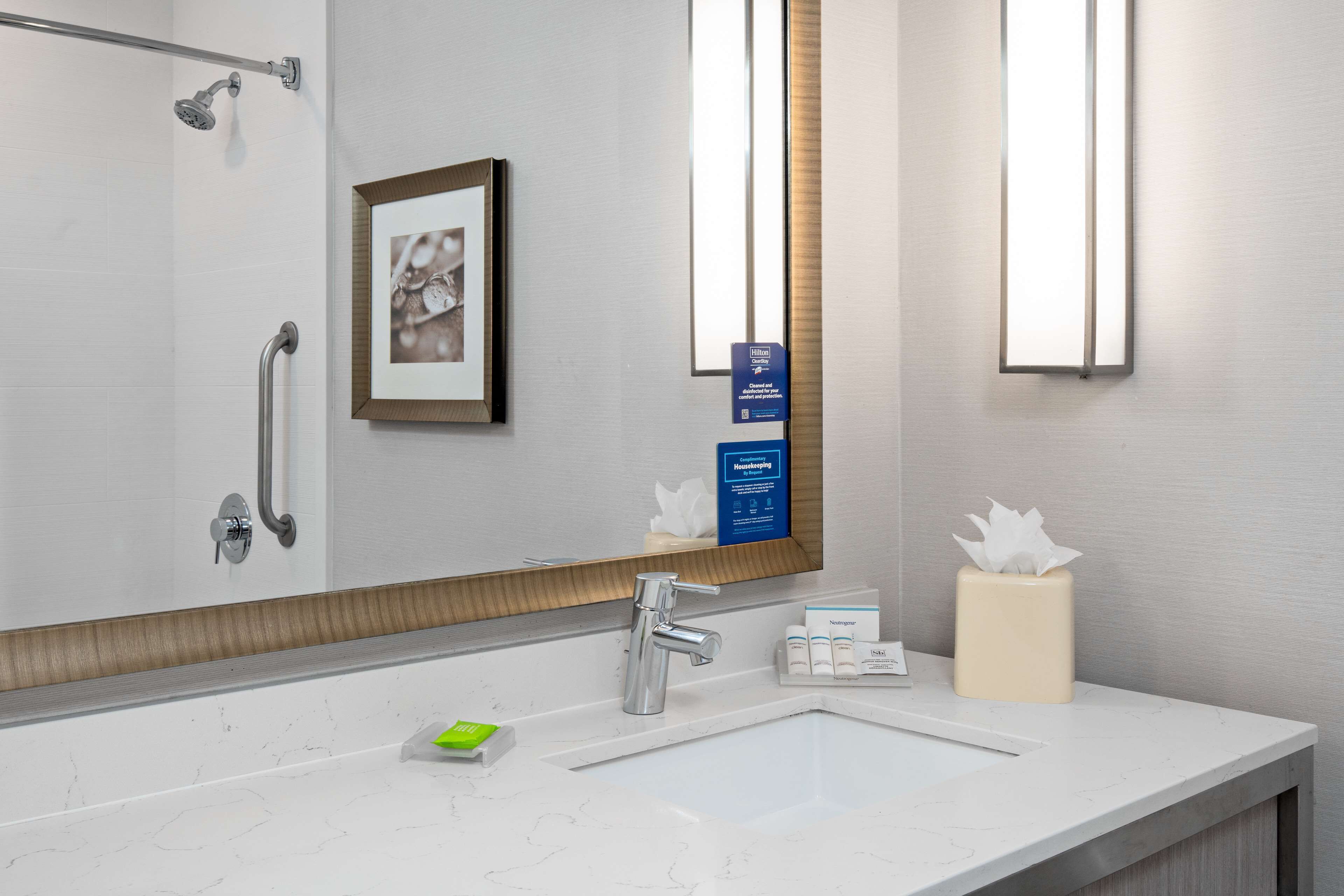 A modern bathroom with a large mirror, a sink with a chrome faucet, a towel dispenser, and a framed picture on the wall.