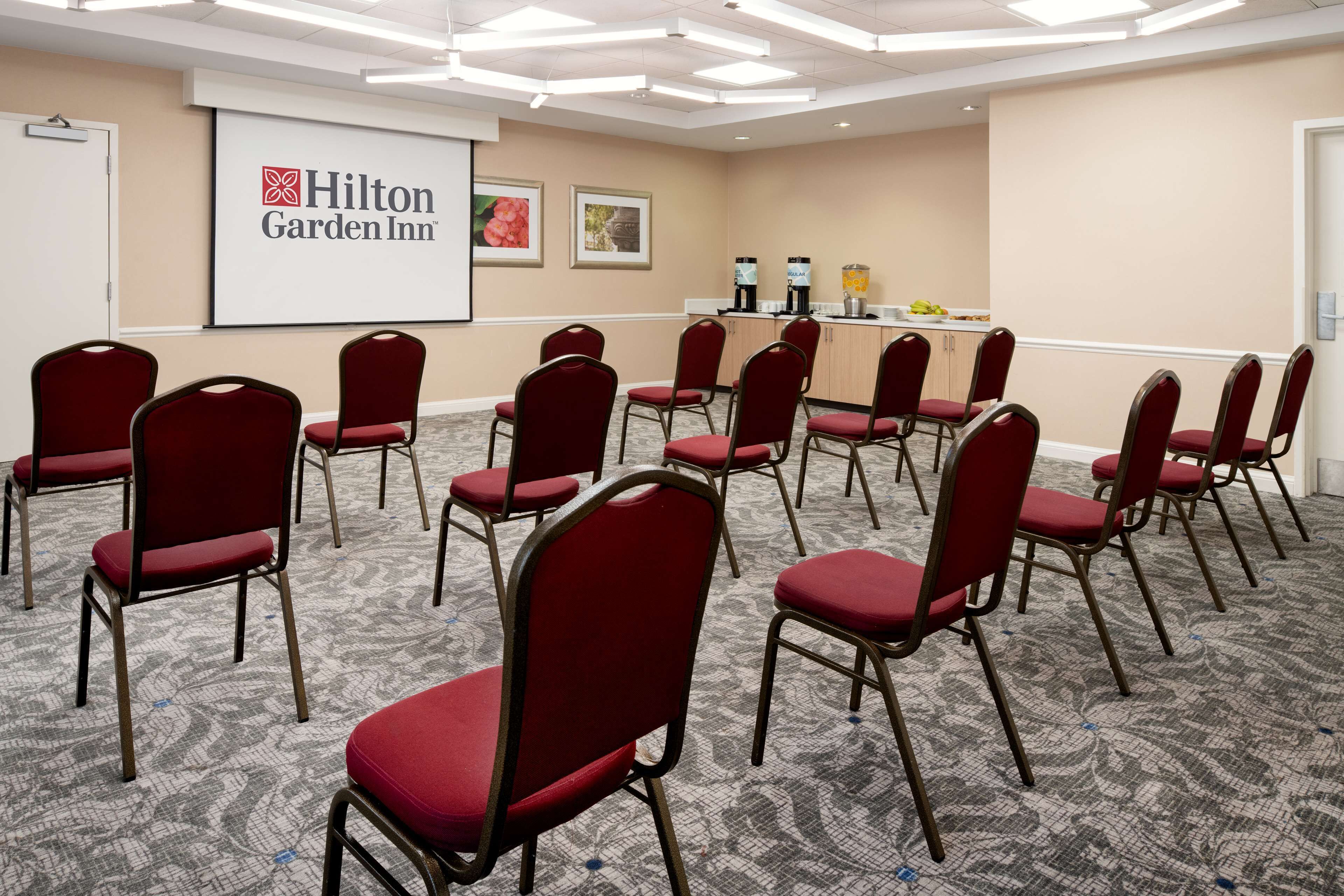 Uma sala de conferência com cadeiras vermelhas dispostas em semicírculo, uma tela de projetor exibindo o logotipo Hilton Garden Inn e um espaço com bebidas e lanches.