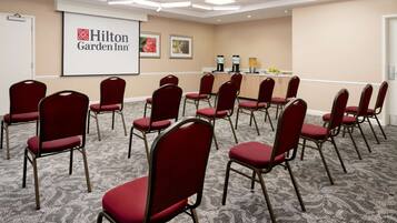 Uma sala de conferência com cadeiras vermelhas dispostas em semicírculo, uma tela de projetor exibindo o logotipo Hilton Garden Inn e um espaço com bebidas e lanches.