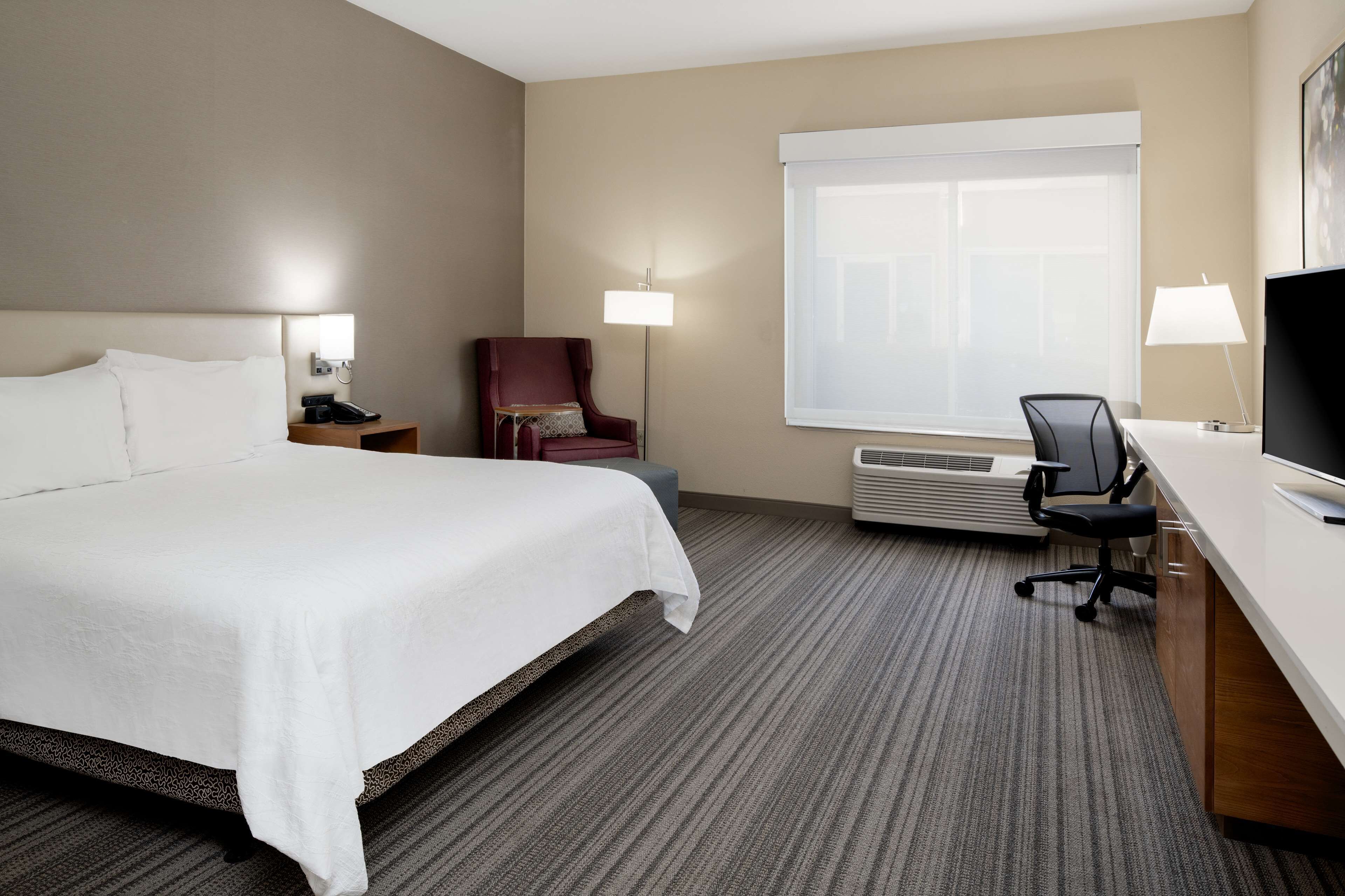 A hotel room with a large bed, a desk with a chair, a TV, and a window with blinds.