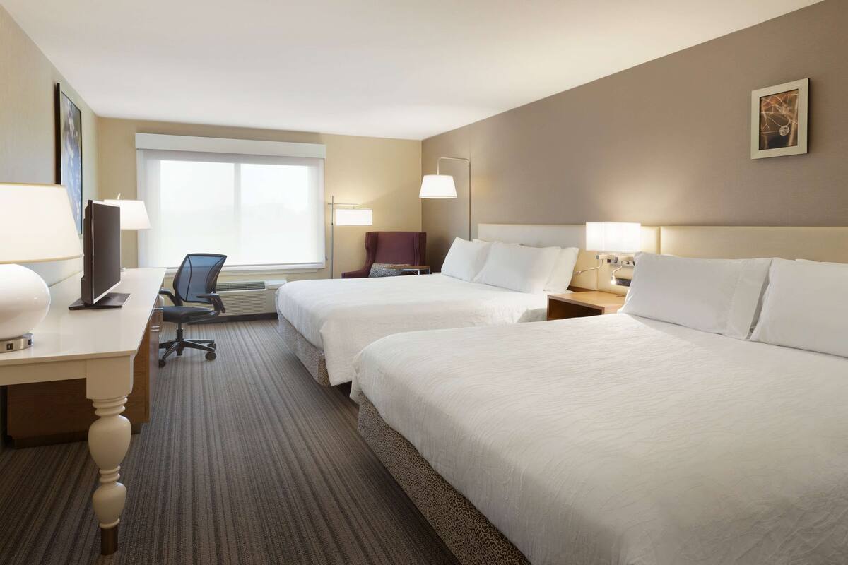 A hotel room with two beds, a desk with a computer, a chair, and a lamp.